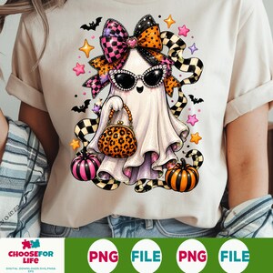 May include: A beige t-shirt featuring a graphic of a white ghost wearing rhinestone-studded sunglasses and a large checkered bow. The ghost holds a leopard-print handbag and is surrounded by small pumpkins, bats, and colorful star shapes.