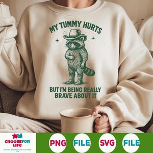 May include: Cream-colored sweatshirt with a green graphic of a raccoon wearing a cowboy hat and boots. The text reads "MY TUMMY HURTS" above the raccoon and "BUT I'M BEING REALLY BRAVE ABOUT IT" below. The sweatshirt is a casual style.