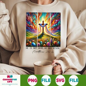 May include: A colourful painting of a cross with a white cloth draped over it. The cross is set against a background of colourful clouds and a green field with Easter eggs. The text "HE IS NOT HERE. HE HAS RISEN. Matthew 28:6" is below the painting.