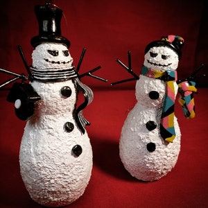 May include: Two white snowman ornaments with black top hats and scarves. One snowman has a black and white striped scarf and the other has a multi-colored scarf. Both snowmen have black eyes and a black mouth.