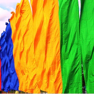 May include: A row of colorful fabric flags waving in the wind. The flags are red, orange, yellow, green, blue, and purple. They are all hanging from poles and are blowing in the breeze.