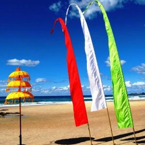 May include: Three colorful fabric flags, red, white, and green, are displayed on a beach with a yellow and orange tiered umbrella in the foreground.