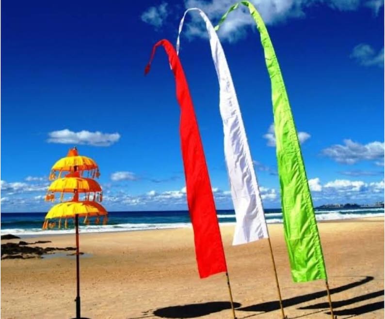 May include: Three colorful fabric flags, red, white, and green, are displayed on a beach with a yellow and orange tiered umbrella in the foreground.