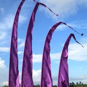 May include: Four purple fabric flags with pointed ends, hanging from bamboo poles against a blue sky. The flags are arranged in a staggered formation, with the largest flag at the back and the smallest flag at the front. The flags are all flowing in the wind.