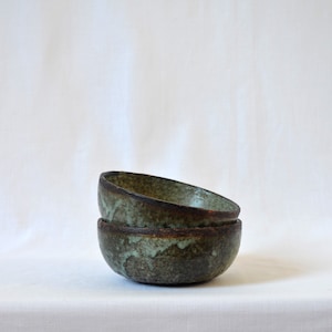 May include: Two stacked ceramic bowls with a green and brown glaze. The bowls are slightly angled and have a rustic, handmade look.