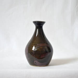 May include: A handcrafted ceramic vase with a dark brown glaze and maroon accents. The vase has a rounded body and a narrow neck, standing approximately 8 inches tall. The surface has a glossy finish, reflecting light. The design is simple and elegant, suitable for displaying flowers.