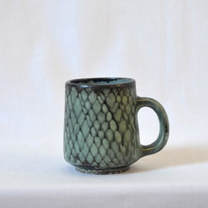 Puede incluir: Una taza de cerámica verde con un patrón texturizado en blanco y negro. La taza tiene una base redondeada y un asa.