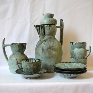 May include: A ceramic tea set in shades of green and gray, featuring a large pitcher, cups, saucers, and small bowls. The set has a unique, handcrafted appearance with dark drips of glaze around the rims.