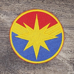 May include: Embroidered patch featuring a circular design with a yellow starburst in the center. The top half is red, the bottom half is blue, and the starburst is outlined in yellow. A yellow border surrounds the entire patch.