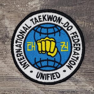 May include: A circular patch featuring the words "INTERNATIONAL TAEKWON-DO FEDERATION" in white around the outer edge. The center has a blue background with a yellow fist and Korean characters. The word "UNIFIED" is at the bottom.