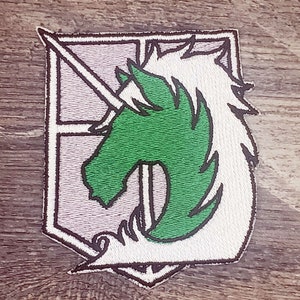 May include: A white and gray embroidered patch with a green unicorn design. The unicorn is facing left and has a horn and mane. The patch has a white border and a gray background.