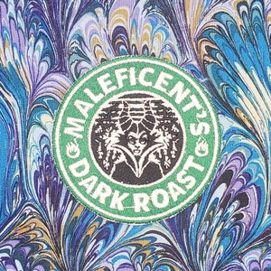 May include: A green and white circular patch with the text "Maleficent's Dark Roast" around the edge. The center of the patch features a black and white illustration of Maleficent from the Disney movie.