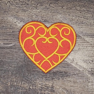 May include: A red heart-shaped patch with a yellow embroidered design. The design features intricate swirls and curves.