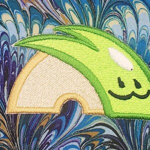 May include: A green and white embroidered patch with a smiling face and a curved shape. The patch is on a blue and brown marbled fabric.