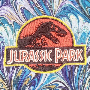 May include: Embroidered patch featuring the Jurassic Park logo. The logo is a red circle with a black silhouette of a dinosaur inside. The text "Jurassic Park" is written in white letters on a black background.