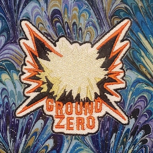 May include: Embroidered patch with a gold center and orange and black lightning bolt design. The patch reads "GROUND ZERO" in orange letters. The background is a blue, purple, and white marbled fabric.