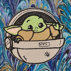 May include: Embroidered patch featuring a green alien-like character in a gray pod. The character has large eyes and pointed ears. The pod is detailed with black stitching and a small hatch. The background has a blue, teal, and gold marbled pattern.