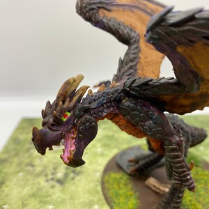 Pode incluir: Uma miniatura de dragão preto e marrom com olhos verdes e boca rosa. O dragão é pintado com uma textura detalhada e está em posição de voo.