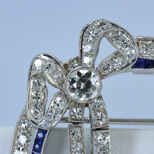 May include: A silver brooch in the shape of a bow with a large, round, clear diamond in the center. The bow is decorated with smaller diamonds and blue sapphires.