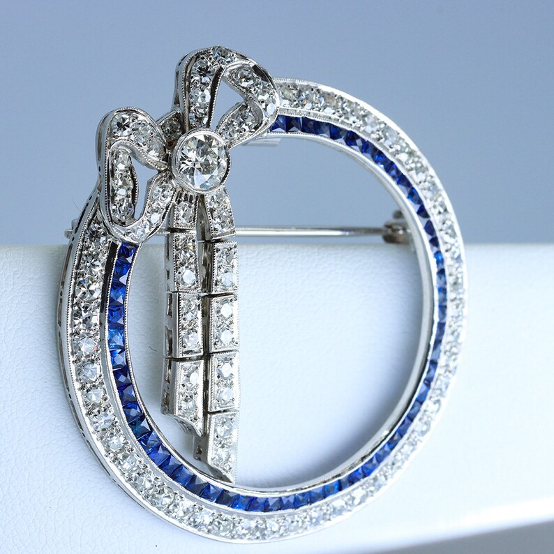 May include: A silver brooch with a diamond and sapphire bow design. The bow is set in a circle of diamonds and sapphires.