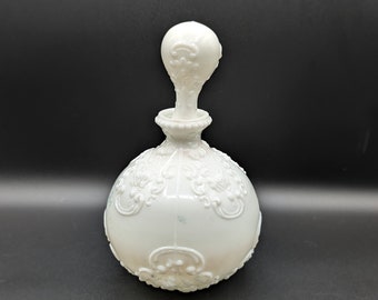 May include: A white milk glass bottle with an ornate design and a round stopper. The bottle has a textured surface and is decorated with raised floral patterns.