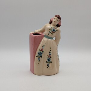 Puede incluir: Una figurita de cerámica de una mujer con un vestido blanco con detalles florales azules y rojos. Está sosteniendo un jarrón rosa con un diseño floral.