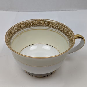 Puede incluir: Una taza de té blanca con un borde dorado y un patrón floral. La taza tiene una pequeña asa y un tazón ancho y poco profundo.