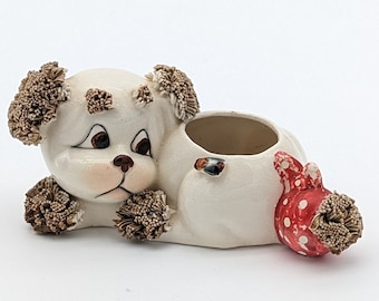 May include: White ceramic dog planter with brown pom-poms for ears and feet. The dog has a red and white polka dot tail.