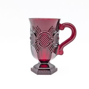 May include: A ruby red glass mug with a decorative pattern. The mug features a handle and sits on a square base. The design includes diamond and circle patterns. The mug is likely used for drinking beverages.