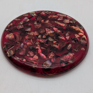 May include: A round, decorative object with a deep red, translucent resin finish. The object is filled with a mosaic of small, irregularly shaped pieces in shades of red, brown, and iridescent colors. The overall effect is a textured, eye-catching design.