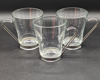 Op de afbeelding: Drie heldere glazen theekopjes met zilverkleurige metalen handvatten en voetjes. De lege kopjes staan in een driehoek op een donkere ondergrond. Geschikt voor thee of koffie.