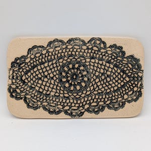 Könnte beinhalten: Ein rechteckiges, beigefarbenes Keramiktablett mit einem dunkelblauen, gehäkelten Deckchen-Design. Das Deckchenmuster ist auf dem Tablett zentriert und weist ein detailliertes, offenes Design auf. Das Tablett hat abgerundete Ecken und eine glatte Oberfläche.