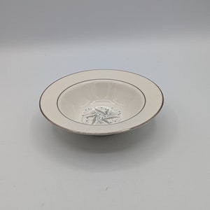 May include: A white ceramic bowl with a gold rim and a blue and grey floral design. The bowl is shallow and has a slightly rounded bottom.