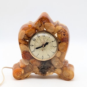 May include: A decorative clock encased in clear resin, featuring a variety of small, light brown stones. The clock face is white with black numerals and hands. The clock is approximately 6 inches tall.