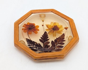 May include: An octagonal wooden frame encases a clear resin display featuring dried flowers. The arrangement includes an orange and a yellow flower, along with dark brown leaves. The frame has a light brown finish.