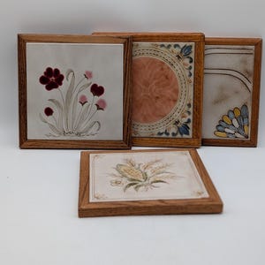 Könnte beinhalten: Set aus vier quadratischen dekorativen Fliesen mit Holzrahmen. Die Fliesen zeigen Blumen- und botanische Designs in Rot-, Rosa-, Gelb- und Blautönen. Die Rahmen sind aus hellbraunem Holz gefertigt.