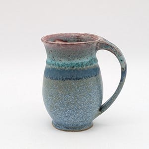 May include: A handmade ceramic mug with a blue, green, and purple glaze. The mug has a wide base and a rounded body with a handle.