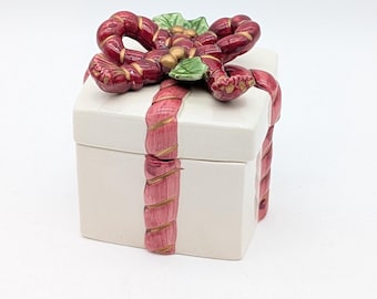 May include: A white ceramic gift box with a red and gold bow and ribbon design. The box is square-shaped and has a lid. The bow features green leaves and gold accents. The ribbon wraps around the box, creating a festive look.