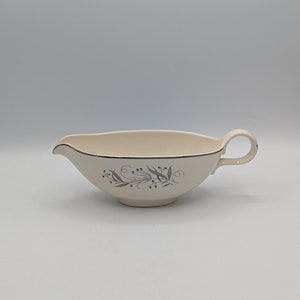 May include: A white ceramic gravy boat with a blue and grey floral design. The boat has a handle on one side and a lip on the other.