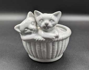 May include: White ceramic planter shaped like a basket with two white ceramic kittens inside. The kittens are sitting in the basket and looking out at the viewer.