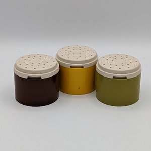 Könnte beinhalten: Drei kleine, runde Salz- und Pfefferstreuer aus Plastik mit weißen Deckeln. Die Streuer sind braun, gelb und grün.
