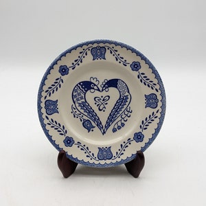 May include: A small, white ceramic plate with a blue and white floral design. The center of the plate features a heart-shaped design with two birds facing each other.