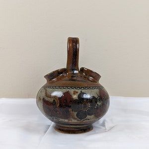 Puede incluir: Una jarra de cerámica marrón con asa y tres picos. La jarra está decorada con un patrón floral en tonos marrones, rojos y azules.