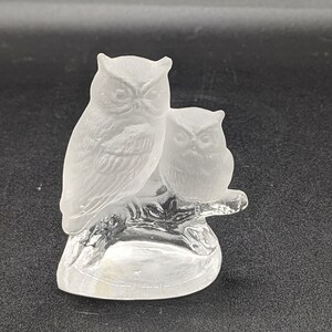 May include: A frosted glass figurine of two owls perched on a branch. The larger owl is on the left and the smaller owl is on the right. The owls are both facing forward with their wings spread out.