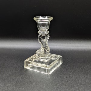 May include: A clear glass candlestick with a square base and a decorative, swirling stem. The candlestick is designed to hold a candle in the top cup.