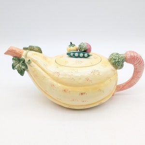 May include: A yellow ceramic teapot shaped like a squash with a pink carrot handle and green leaves. The lid has a green pea pod with green peas and a small red tomato and a purple turnip on top.