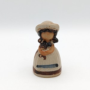 May include: A ceramic figurine of a girl wearing a white hat and brown dress holding a bouquet of flowers. The figurine is painted in brown, white, and blue.
