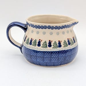 Puede incluir: Jarra de cerámica con asa y boquilla azules. La jarra presenta un estampado de lunares azules y blancos en la parte inferior, con una banda de muñecos de nieve y árboles de Navidad. El borde superior tiene un diseño de copo de nieve.