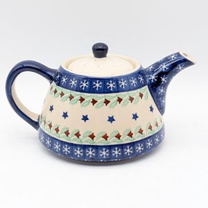 Puede incluir: Una tetera de cerámica con asa, boquilla y borde azules. El cuerpo es de color crema con un patrón de copos de nieve azules, hojas verdes y estrellas rojas. La tapa es de color crema con un pomo azul oscuro.