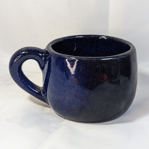 Könnte beinhalten: Dunkelblaue Keramiktasse mit abgerundeter Form und Henkel. Die Tasse hat eine glänzende Oberfläche und einen dunkelblauen Innenraum. Geeignet für Heißgetränke wie Tee oder Kaffee. Durchmesser ca. 10 cm.